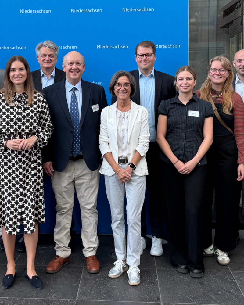 Vor einem blauen Hintergrund mit dem Logo der Landesvertretung Niedersachsen steht eine Gruppe von Menschen in Businesskleidung und lächelt in die Kamera. Das Foto wirkt offiziell und wurde vermutlich im Rahmen eines parlamentarischen Frühstücks oder einer politischen Veranstaltung aufgenommen.