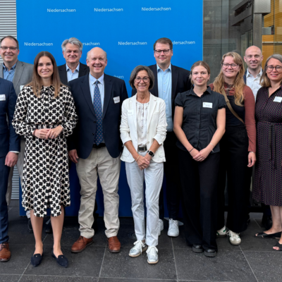 Vor einem blauen Hintergrund mit dem Logo der Landesvertretung Niedersachsen steht eine Gruppe von Menschen in Businesskleidung und lächelt in die Kamera. Das Foto wirkt offiziell und wurde vermutlich im Rahmen eines parlamentarischen Frühstücks oder einer politischen Veranstaltung aufgenommen.