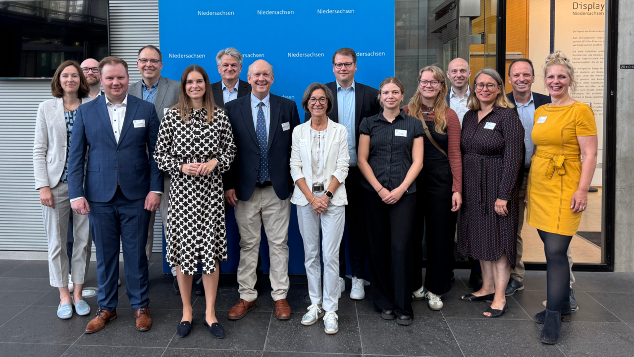 Vor einem blauen Hintergrund mit dem Logo der Landesvertretung Niedersachsen steht eine Gruppe von Menschen in Businesskleidung und lächelt in die Kamera. Das Foto wirkt offiziell und wurde vermutlich im Rahmen eines parlamentarischen Frühstücks oder einer politischen Veranstaltung aufgenommen.