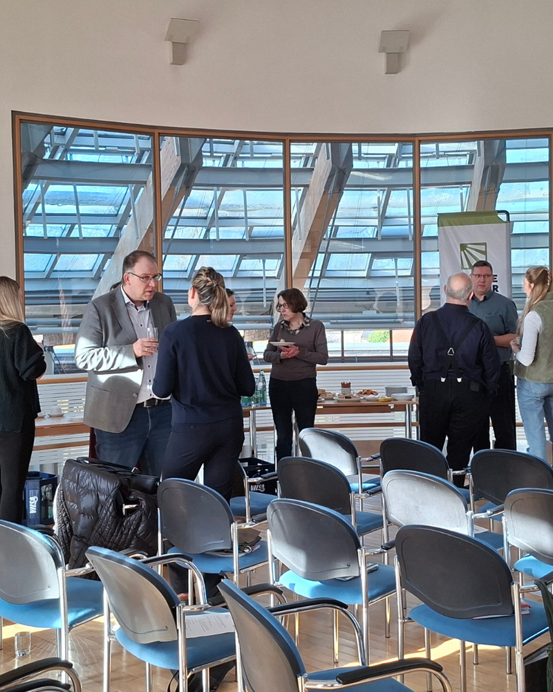 Mehrere Personen stehen in kleinen Gruppen in einem hellen Seminarraum und unterhalten sich während einer Pause; im Vordergrund sind leere Reihenstühle zu sehen. Rechts gestikuliert eine Frau neben einer Leinwand, während links andere Teilnehmende Getränke und Snacks vom Tisch nehmen.