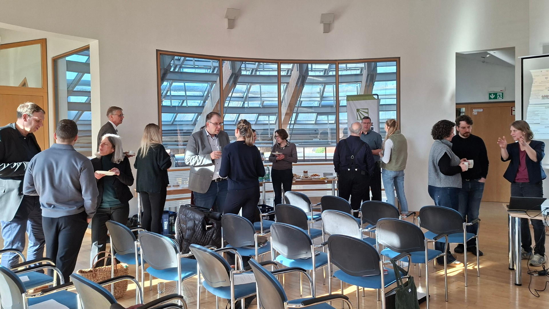 Mehrere Personen stehen in kleinen Gruppen in einem hellen Seminarraum und unterhalten sich während einer Pause; im Vordergrund sind leere Reihenstühle zu sehen. Rechts gestikuliert eine Frau neben einer Leinwand, während links andere Teilnehmende Getränke und Snacks vom Tisch nehmen.