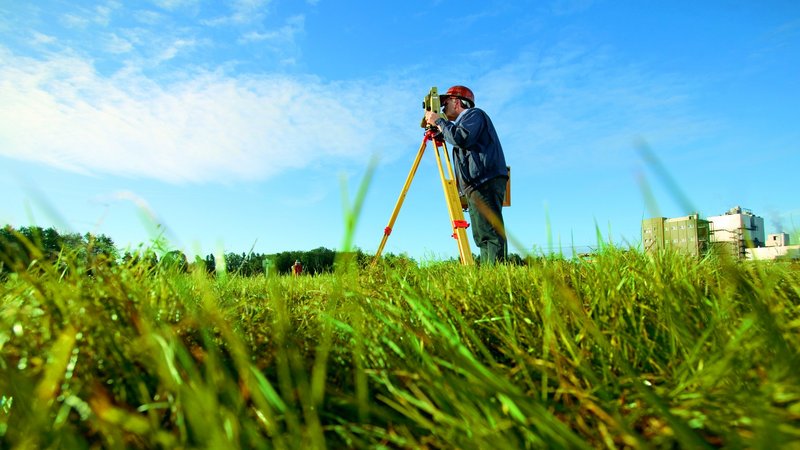 Diese Aufnahme zeigt einen Vermessungsingenieur, der mit einem Theodoliten an einer Baustelle arbeitet. Inmitten einer üppigen Wiese mit hohem Gras steht der Mann konzentriert an seinem Messgerät, das er zur Vermessung und Kartierung des Geländes einsetzt.