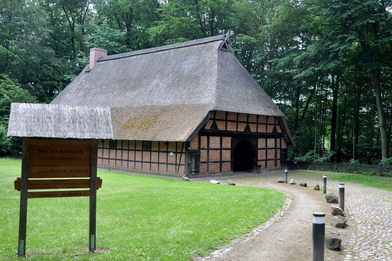 Das Bild zeigt ein historisches Bauernhaus mit Reetdach, umgeben von einem grünen Park und einem Waldstück. Die traditionelle, halbholzverkleidete Architektur in Kombination mit der idyllischen Naturumgebung verleiht der Szene einen authentischen, ländlichen Charakter.