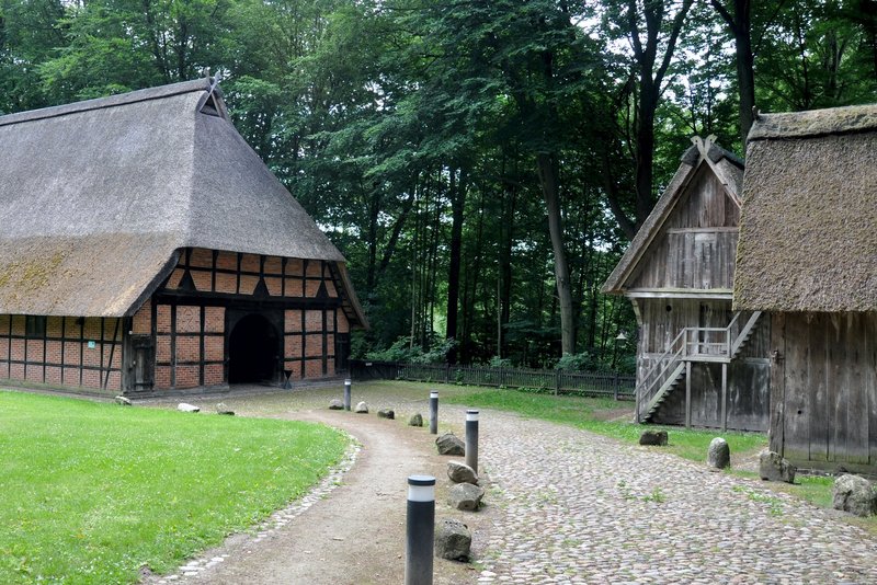 Das Bild zeigt ein historisches Bauernhaus mit Reetdach, umgeben von einem grünen Park und einem Waldstück. Die traditionelle, halbholzverkleidete Architektur in Kombination mit der idyllischen Naturumgebung verleiht der Szene einen authentischen, ländlichen Charakter.