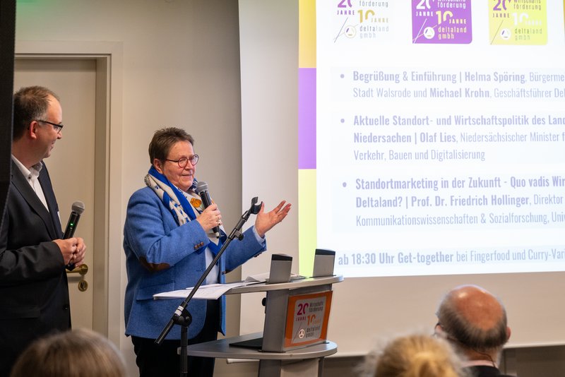 Auf dem Bild stehen ein Mann und eine Frau an einem Stehpult, beide halten ein Mikrofon hält und der Mann spricht. Vor ihnen sitzt ein Publikum, und das Rednerpult trägt ein Schild mit der Aufschrift „20 Jahre Wirtschaftsförderung Deltaland GmbH“. Im Hintergrund ist eine Projektion zu sehen.