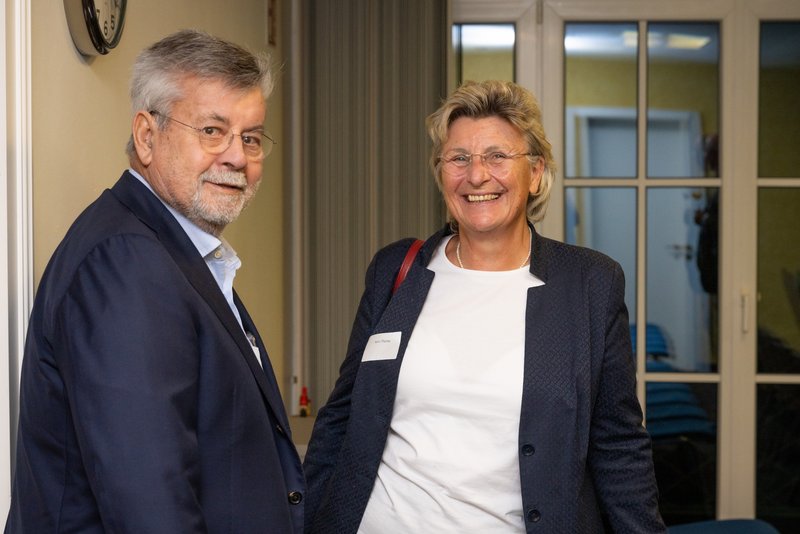 Auf dem Bild lächeln zwei Personen freundlich in die Kamera, während sie in einem hellen Raum stehen. Der Mann trägt einen dunklen Anzug und die Frau eine hellere Jacke, beide scheinen sich in einer geselligen Atmosphäre zu befinden.