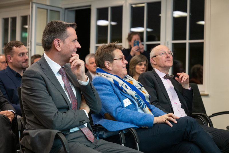 Das Bild zeigt eine Gruppe von Personen, die in der ersten Reihe einer Veranstaltung aufmerksam nach vorne blicken und offenbar einem Vortrag oder einer Präsentation folgen. Im Vordergrund sitzen drei Personen, darunter eine Frau im blauen Blazer, flankiert von zwei Männern in Anzug und Krawatte bzw. Fliege, während im Hintergrund weitere Zuhörer und eine fotografierende Person zu sehen sind.