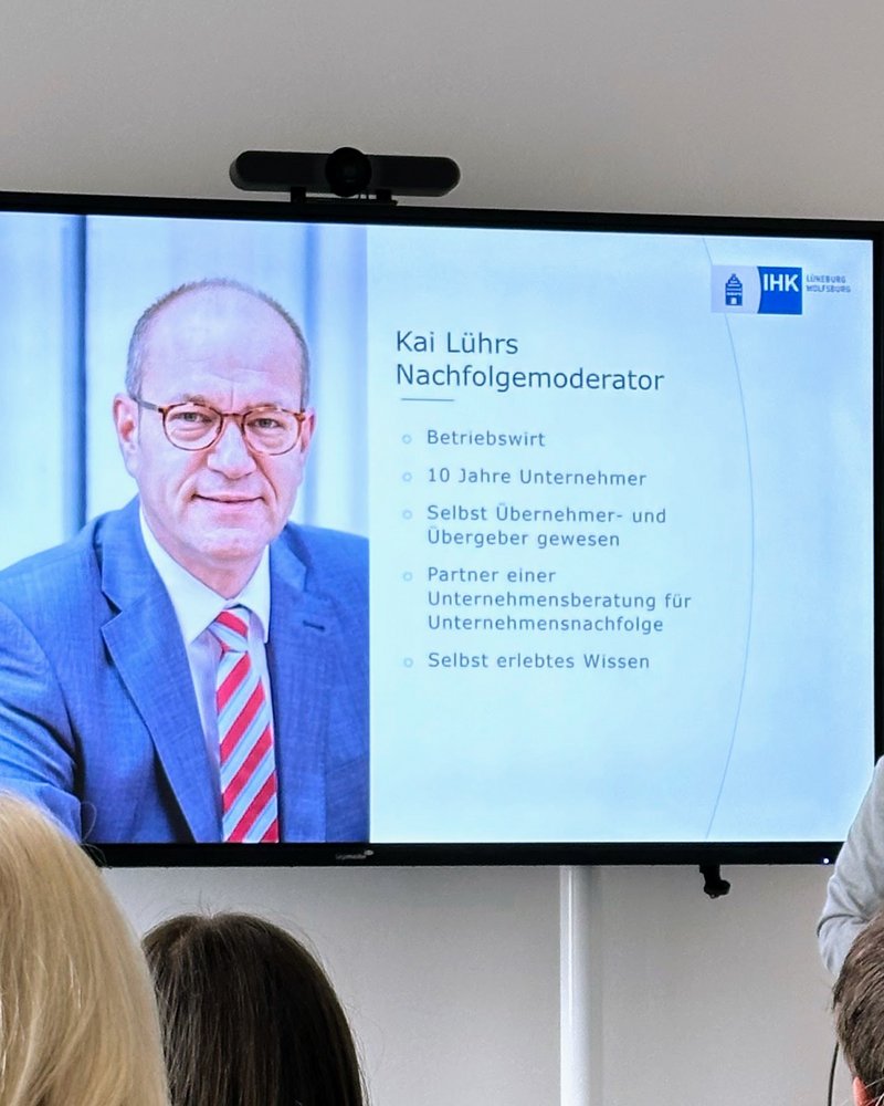 IHK-Nachfolgemoderator Kai Lührs (Mann mit Brille in hellgrauem Sakko) hält einen Vortrag. Neben im ist ein großer Monitor zu sehen, der die aktuelle Vortragsfolie von Kai Lührs darstellt (hier ein paar Infos zur Person Lührs selbst).