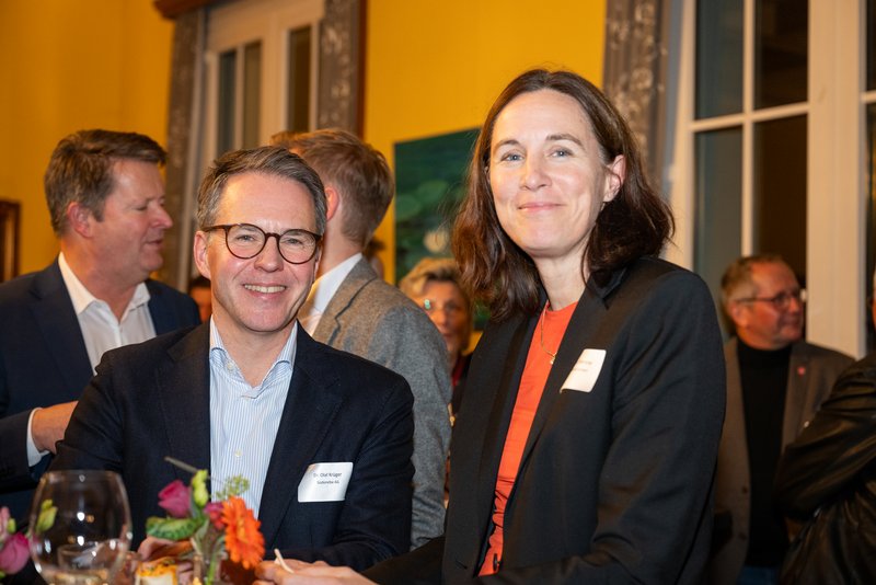 Das Bild zeigt eine gesellige Veranstaltung, bei der eine Gruppe von Menschen miteinander interagiert. Im Vordergrund lächeln ein Mann mit Brille und eine Frau, die sich freundlich unterhalten, während im Hintergrund weitere Gäste zu sehen sind.
