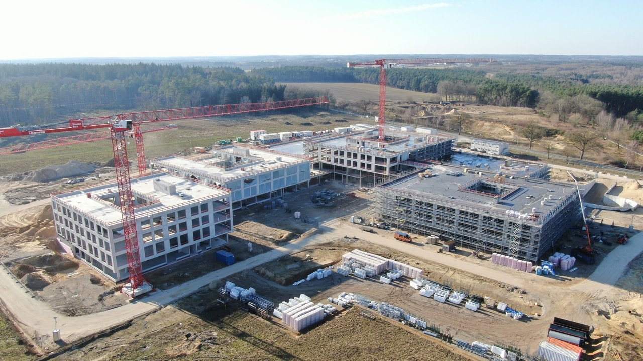 Das Luftbild zeigt eine große Baustelle mit mehreren mehrstöckigen Gebäuden im Rohbau, die von zwei markanten roten Liebherr-Turmdrehkranen überragt werden. Das weitläufige Gelände ist umgeben von Waldgebieten und offenen Feldflächen, während auf dem Baufeld zahlreiche Baumaterialien und Baufahrzeuge zu erkennen sind.