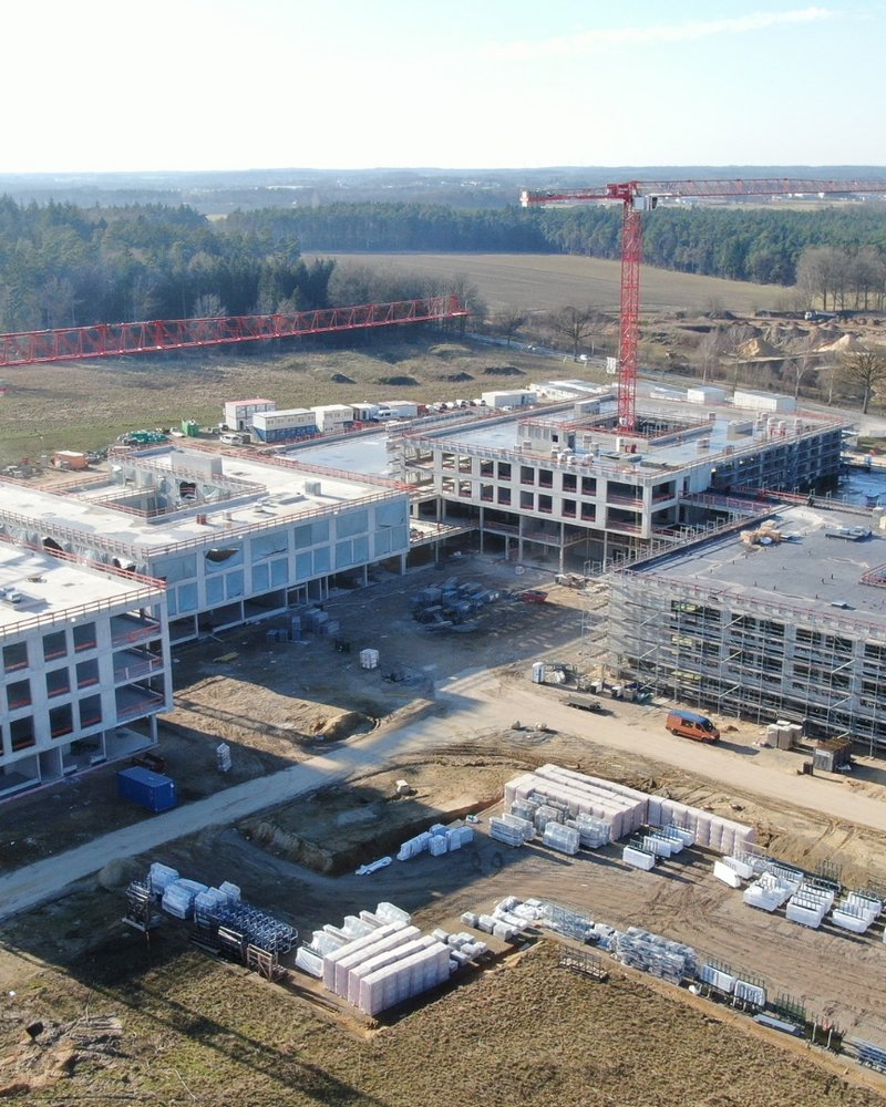Das Luftbild zeigt eine große Baustelle mit mehreren mehrstöckigen Gebäuden im Rohbau, die von zwei markanten roten Liebherr-Turmdrehkranen überragt werden. Das weitläufige Gelände ist umgeben von Waldgebieten und offenen Feldflächen, während auf dem Baufeld zahlreiche Baumaterialien und Baufahrzeuge zu erkennen sind.