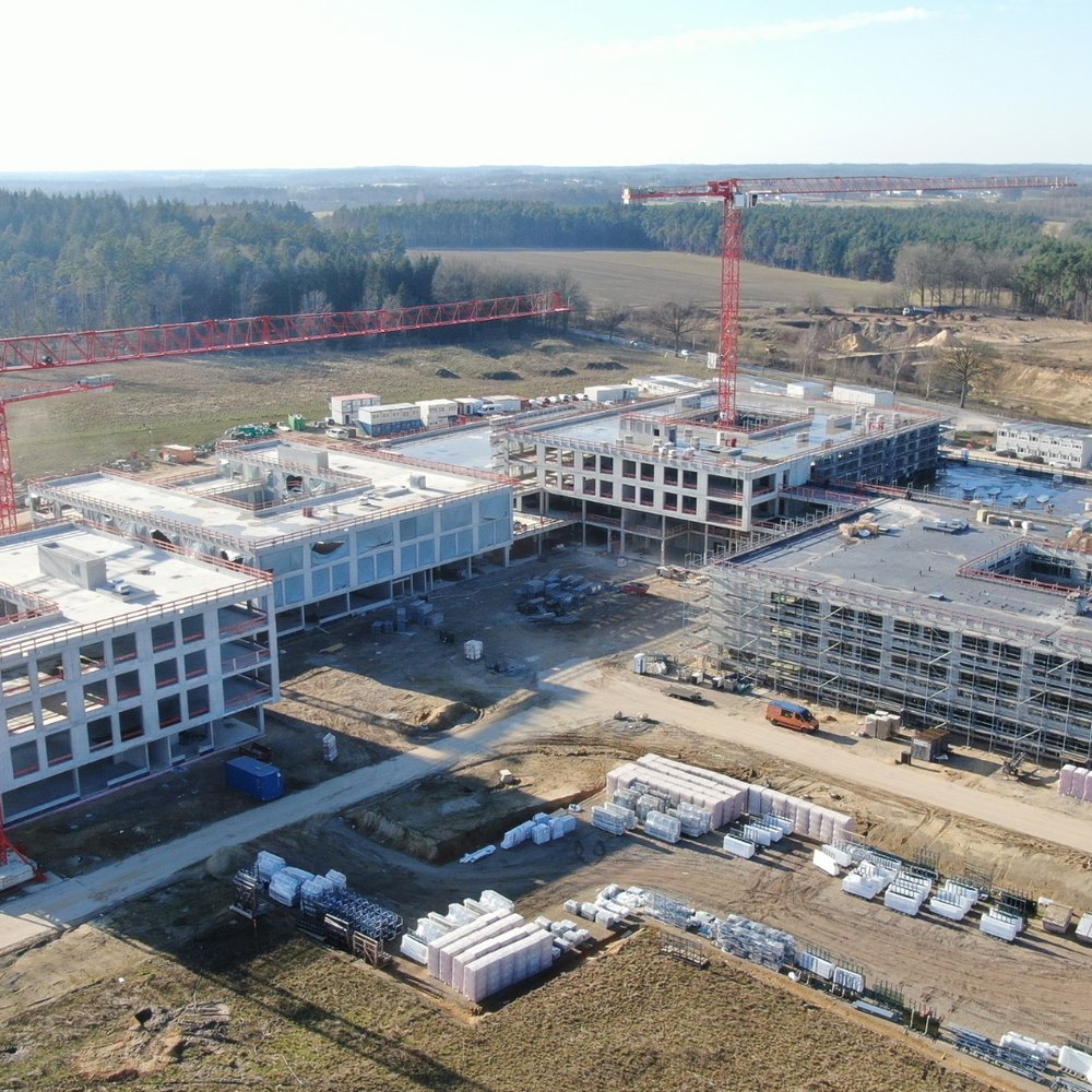 Das Luftbild zeigt eine große Baustelle mit mehreren mehrstöckigen Gebäuden im Rohbau, die von zwei markanten roten Liebherr-Turmdrehkranen überragt werden. Das weitläufige Gelände ist umgeben von Waldgebieten und offenen Feldflächen, während auf dem Baufeld zahlreiche Baumaterialien und Baufahrzeuge zu erkennen sind.