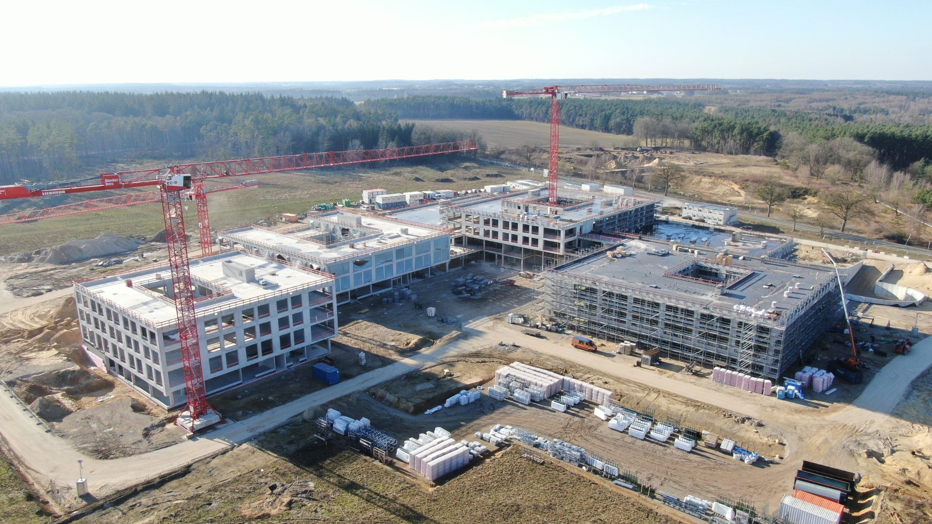 Das Luftbild zeigt eine große Baustelle mit mehreren mehrstöckigen Gebäuden im Rohbau, die von zwei markanten roten Liebherr-Turmdrehkranen überragt werden. Das weitläufige Gelände ist umgeben von Waldgebieten und offenen Feldflächen, während auf dem Baufeld zahlreiche Baumaterialien und Baufahrzeuge zu erkennen sind.