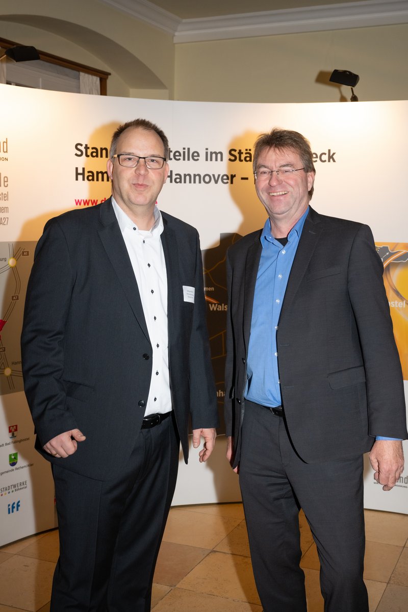 Das Bild zeigt zwei Männer, die freundlich vor einem Banner mit der Aufschrift "deltaland" stehen. Sie tragen formelle Kleidung und scheinen an einer geschäftlichen Veranstaltung teilzunehmen.