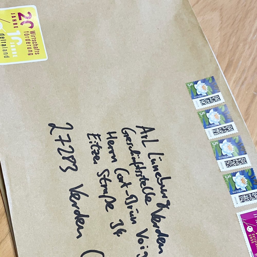Das Bild zeigt einen braunen Briefumschlag mit handschriftlicher Adresse. Er ist mit mehreren Briefmarken frankiert. Oben links befindet sich ein gelber Aufkleber mit einem Jubiläumshinweis. Der Umschlag liegt auf einer Holzoberfläche.