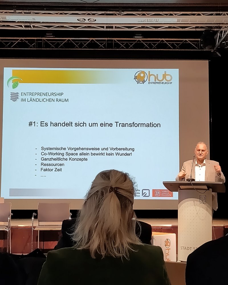 Das Bild zeigt einen Redner auf einer Bühne, der vor einem Publikum einen Vortrag hält. Auf der Leinwand hinter ihm steht der Titel "#1: Es handelt sich um eine Transformation" mit weiteren Stichpunkten zur systemischen Vorgehensweise, ganzheitlichen Konzepten, Ressourcen und dem Faktor Zeit.