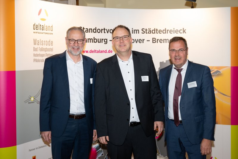 Das Bild zeigt drei Männer, die vor einem großen Banner mit dem Logo von DeltaLand stehen. Sie lächeln und tragen formelle Kleidung, während sie an einer Veranstaltung teilnehmen.