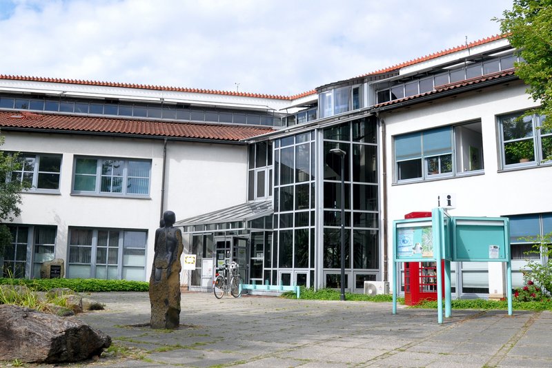 Das Bild zeigt ein modernes Gebäude mit einer interessanten Architektur, das offensichtlich als öffentliche Einrichtung genutzt wird. Der Außenbereich ist mit Kunstwerken, Grünflächen und Fahrradständern gestaltet, die auf einen Bildungskontext hinweisen.