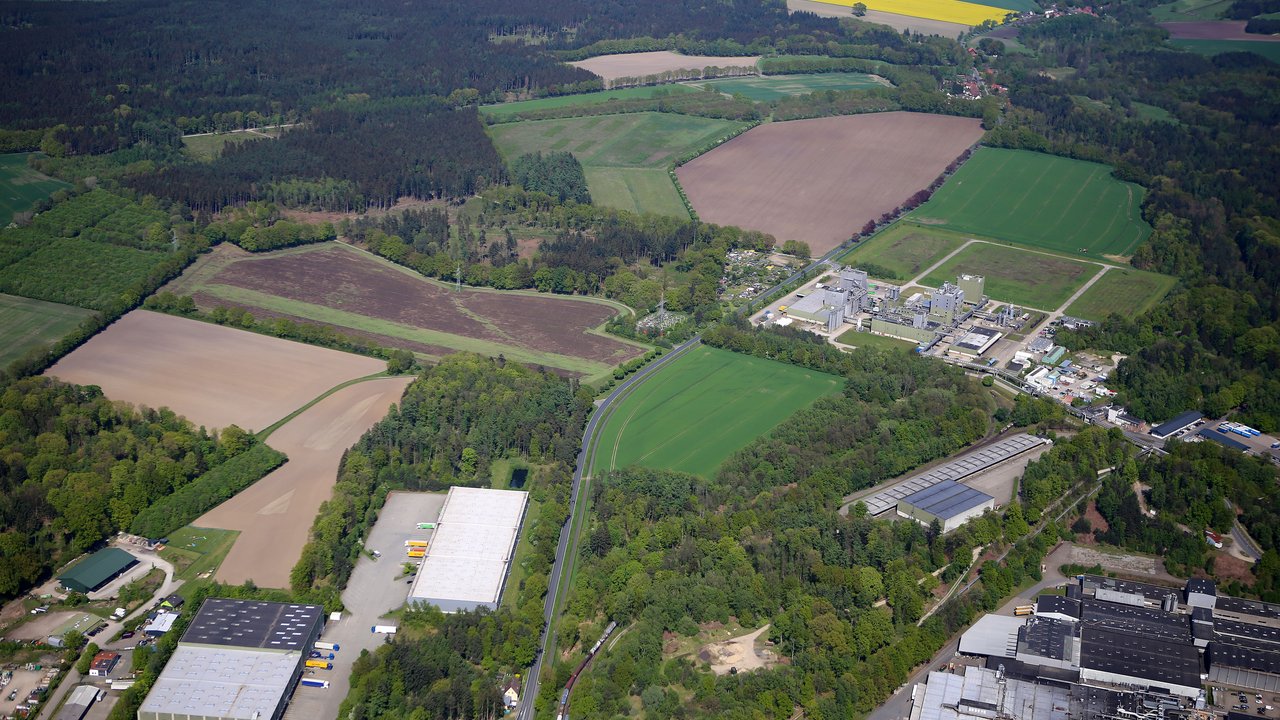 Diese Luftaufnahme zeigt eine gemischte Landschaft aus industriellen Anlagen, Feldern, Wäldern und kleinen Siedlungen. Das Bild gibt einen Überblick über eine Region, in der Industrie und Landwirtschaft nebeneinander existieren und eine abwechslungsreiche Umgebung mit ländlichen und urbanen Elementen bilden.