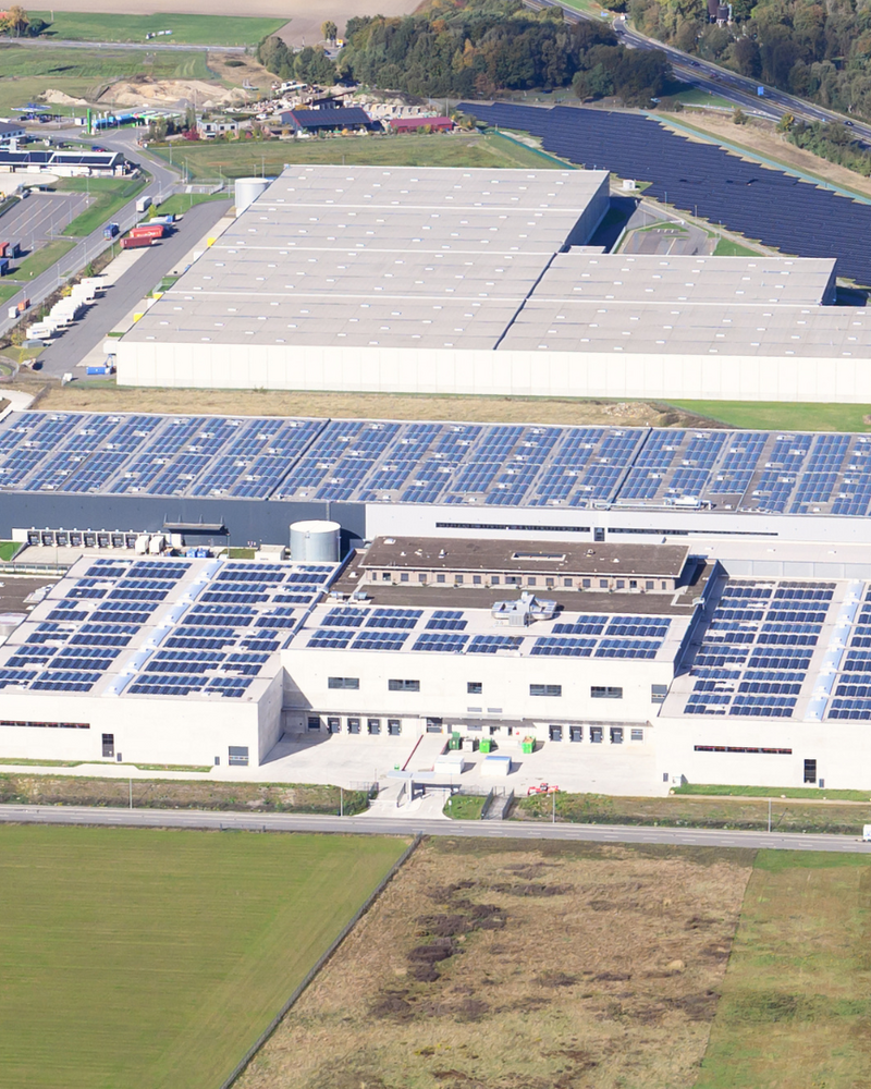 Das Bild zeigt eine Luftaufnahme einer großen Halle, die mit Solarzellen auf dem Dach ausgestattet ist, umgeben von weitläufigen Grünflächen und weiteren Lagerhallen. Die Landschaft im Hintergrund ist von Bäumen und Feldern geprägt.