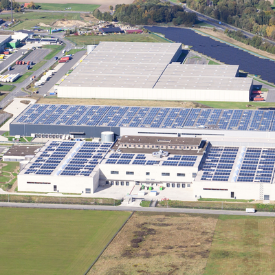Das Bild zeigt eine Luftaufnahme einer großen Halle, die mit Solarzellen auf dem Dach ausgestattet ist, umgeben von weitläufigen Grünflächen und weiteren Lagerhallen. Die Landschaft im Hintergrund ist von Bäumen und Feldern geprägt.