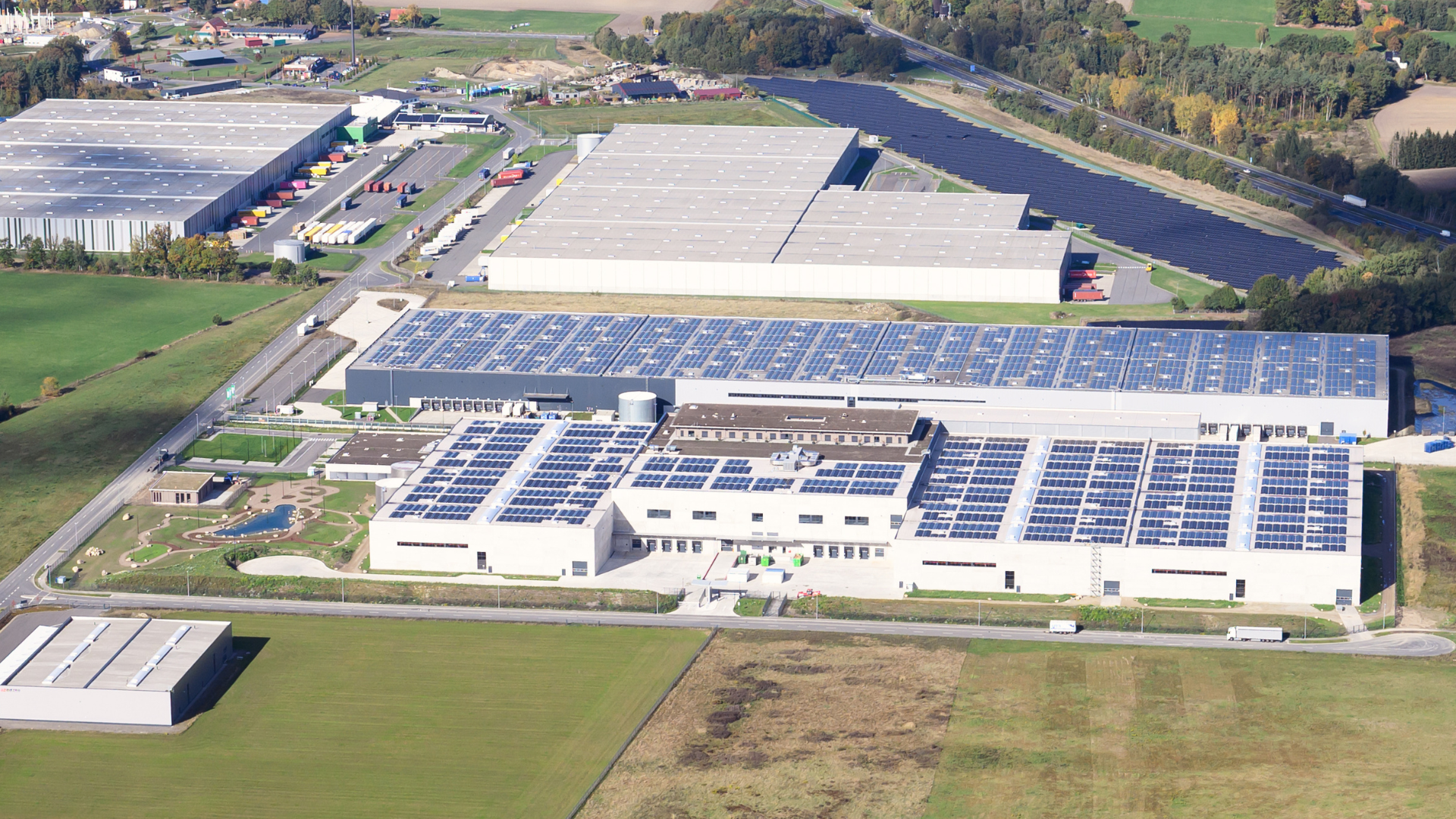 Das Bild zeigt eine Luftaufnahme einer großen Halle, die mit Solarzellen auf dem Dach ausgestattet ist, umgeben von weitläufigen Grünflächen und weiteren Lagerhallen. Die Landschaft im Hintergrund ist von Bäumen und Feldern geprägt.