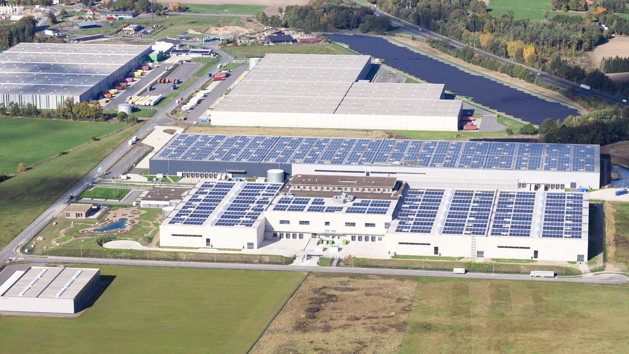 | Ⓒ Deltaland Das Bild zeigt eine Luftaufnahme einer großen Halle, die mit Solarzellen auf dem Dach ausgestattet ist, umgeben von weitläufigen Grünflächen und weiteren Lagerhallen. Die Landschaft im Hintergrund ist von Bäumen und Feldern geprägt.