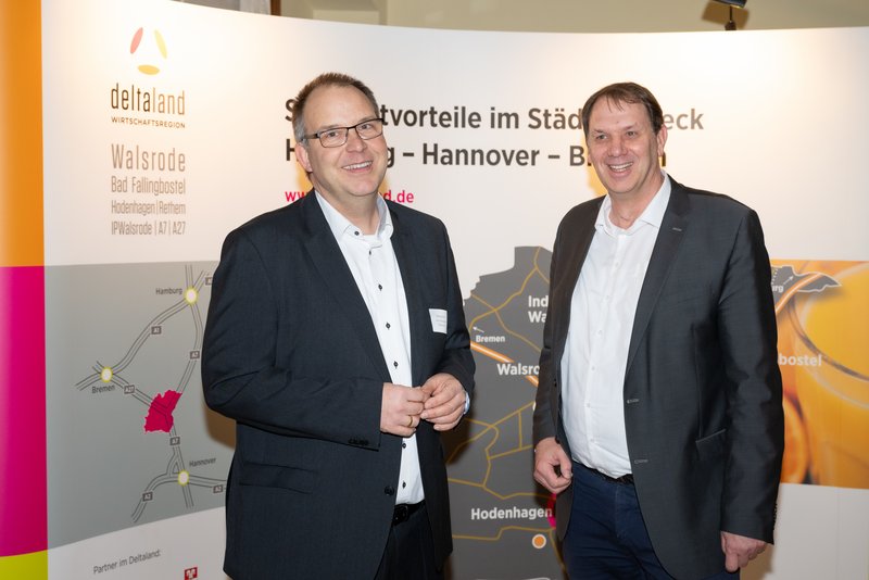 Das Bild zeigt zwei Männer, die freundlich vor einem Banner mit der Aufschrift "deltaland" stehen. Sie tragen formelle Kleidung und scheinen an einer geschäftlichen Veranstaltung teilzunehmen.