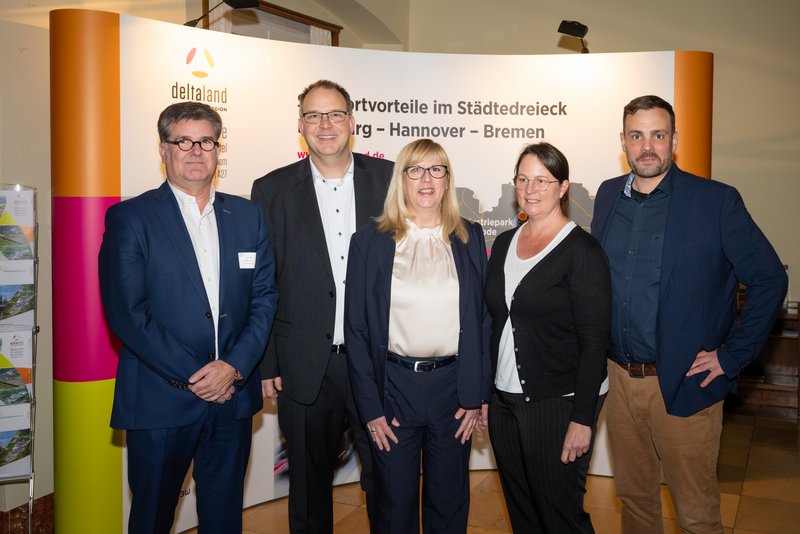 Das Bild zeigt eine Gruppe von fünf Personen, die lächelnd vor einem großen Banner mit der Aufschrift "deltaland" stehen. Die Anwesenden sind in formeller Kleidung und scheinen an einer Veranstaltung teilzunehmen.
