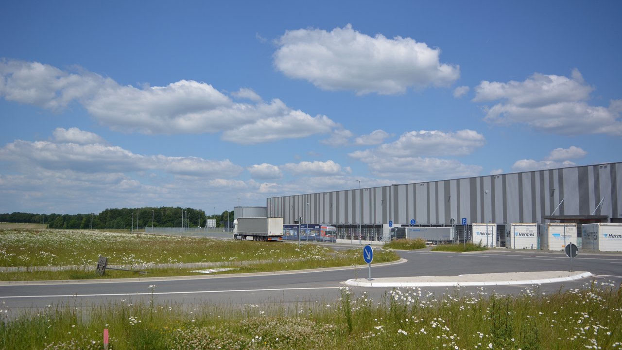  Das Bild zeigt eine große Logistikanlage mit mehreren Lagerhallen und Lkw-Stellplätzen. Umgeben ist das Gelände von einer üppigen Blumenwiese mit Gräsern und Wildblumen, die einen schönen Kontrast zu den grauen Industriegebäuden bildet.