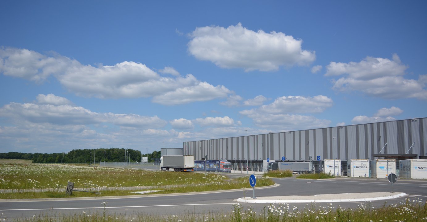  Das Bild zeigt eine große Logistikanlage mit mehreren Lagerhallen und Lkw-Stellplätzen. Umgeben ist das Gelände von einer üppigen Blumenwiese mit Gräsern und Wildblumen, die einen schönen Kontrast zu den grauen Industriegebäuden bildet.