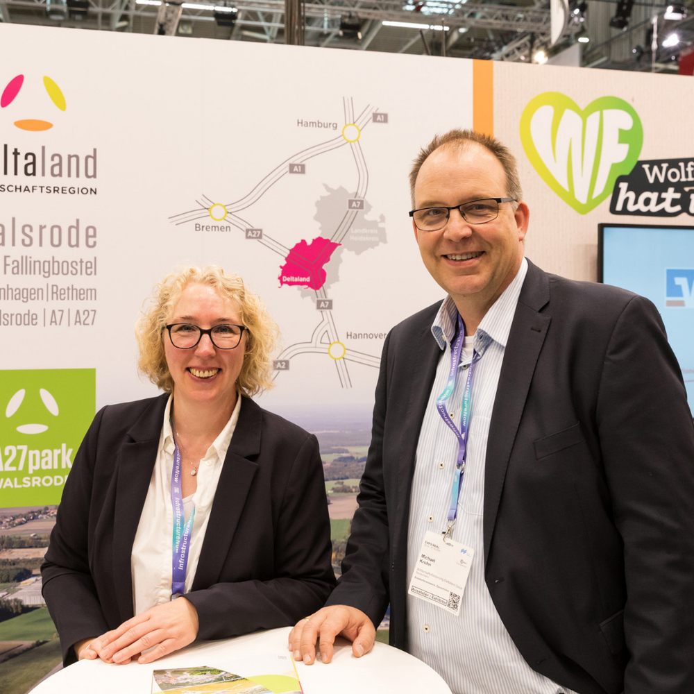 Eine Frau mit lockigen blonden Haaren und ein Mann mit Brille stehen lächelnd an einem Stehtisch am Messestand der Deltaland Wirtschaftsregion. Im Hintergrund sind das Deltaland-Logo, eine Anfahrtskarte und der A27park Walsrode zu sehen.