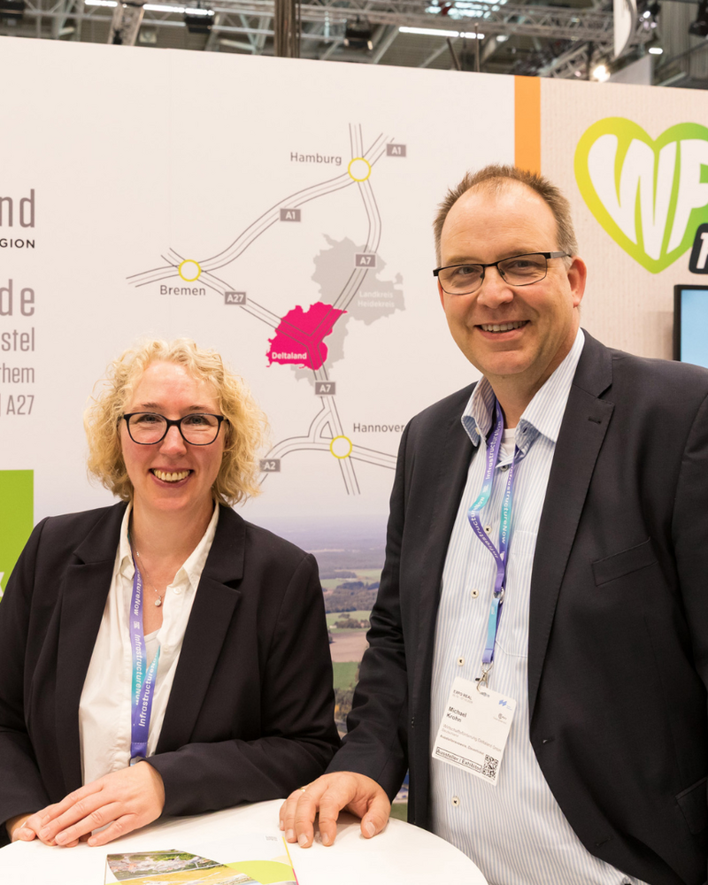 Eine Frau mit lockigen blonden Haaren und ein Mann mit Brille stehen lächelnd an einem Stehtisch am Messestand der Deltaland Wirtschaftsregion. Im Hintergrund sind das Deltaland-Logo, eine Anfahrtskarte und der A27park Walsrode zu sehen.