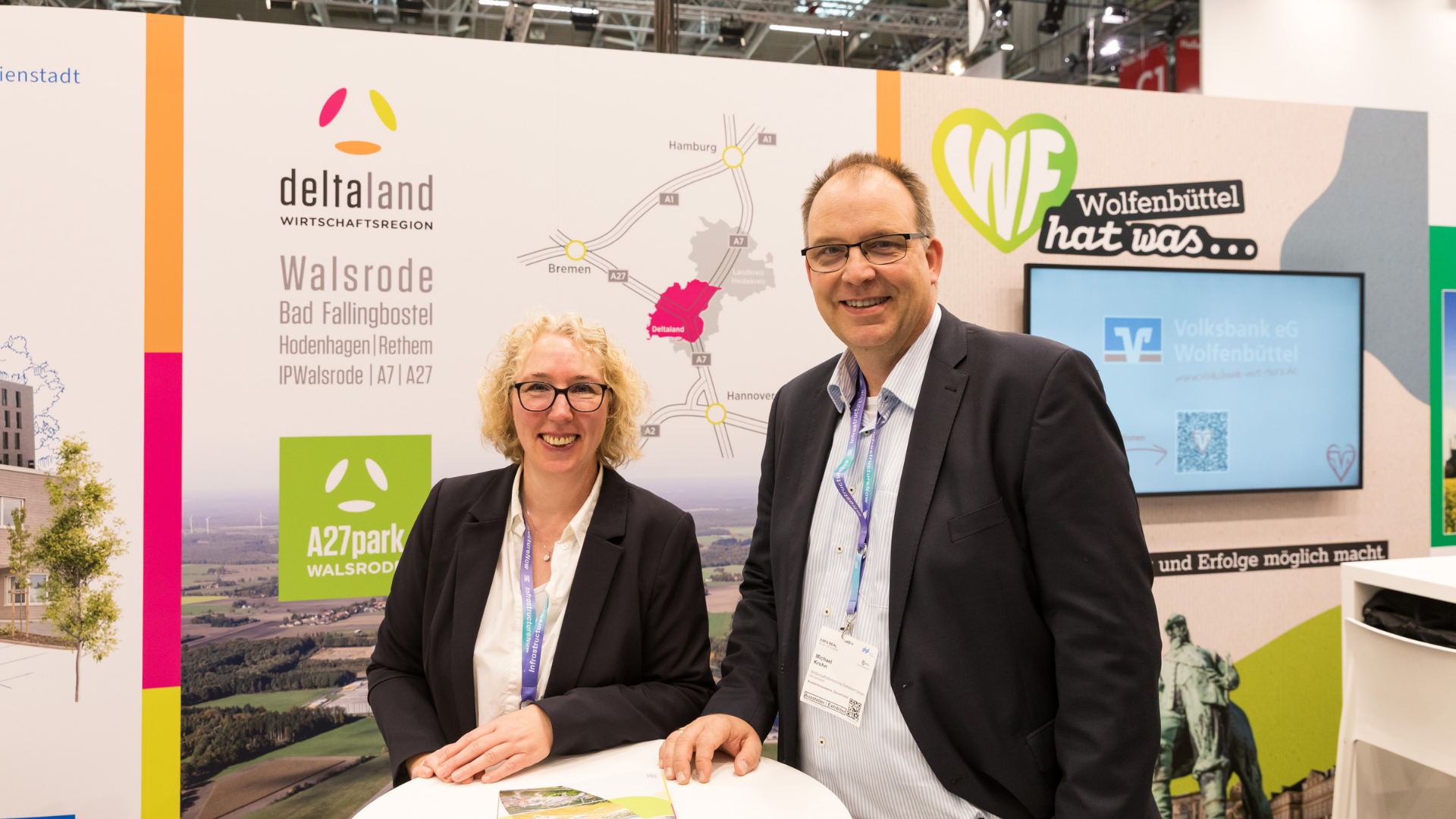 Eine Frau mit lockigen blonden Haaren und ein Mann mit Brille stehen lächelnd an einem Stehtisch am Messestand der Deltaland Wirtschaftsregion. Im Hintergrund sind das Deltaland-Logo, eine Anfahrtskarte und der A27park Walsrode zu sehen.