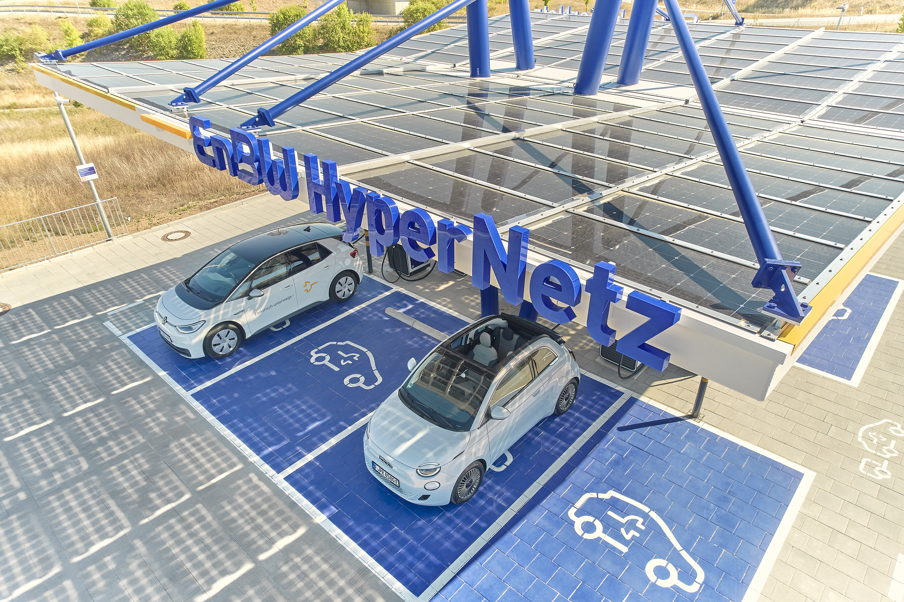 Das Bild zeigt eine große Photovoltaik-Anlage, die ein Parkplatz für Elektrofahrzeuge überdacht. Auf den Solarpaneelen sind zwei weiße Elektroautos geparkt, die offensichtlich an der Ladestation aufgeladen werden. Die Anlage vermittelt einen modernen, nachhaltigen Eindruck und bietet Autofahrern die Möglichkeit, ihre Fahrzeuge umweltfreundlich aufzuladen.