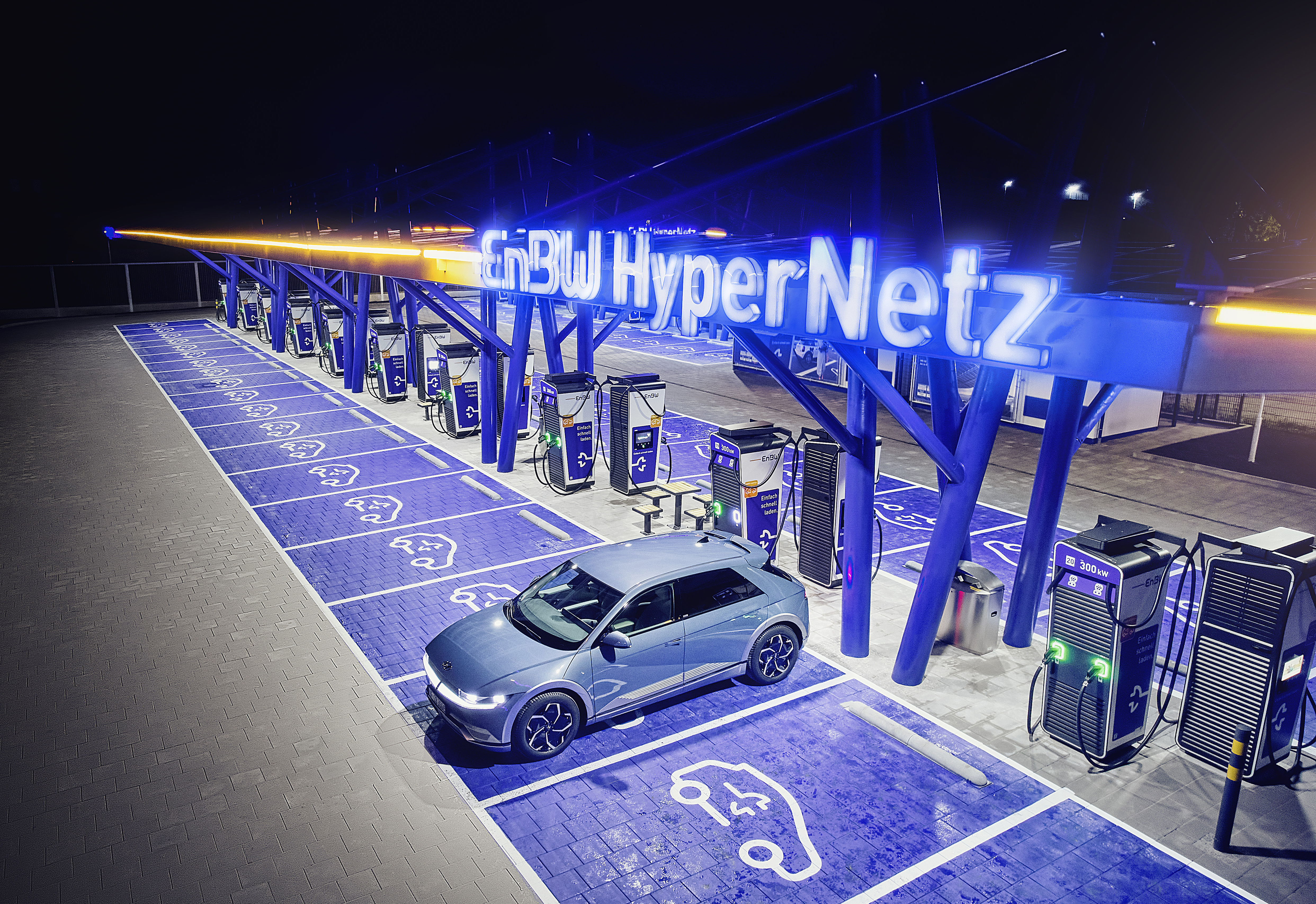 Das Bild zeigt eine Elektroauto-Ladestation mit mehreren Ladepunkten, die von einer großen Photovoltaik-Überdachung beschattet werden. Im Vordergrund steht ein silbernes Elektrofahrzeug an einem der Ladepunkte. Die gesamte Anlage ist in blauen und orangefarbenen Tönen gestaltet und vermittelt einen modernen, hochtech-Charakter.