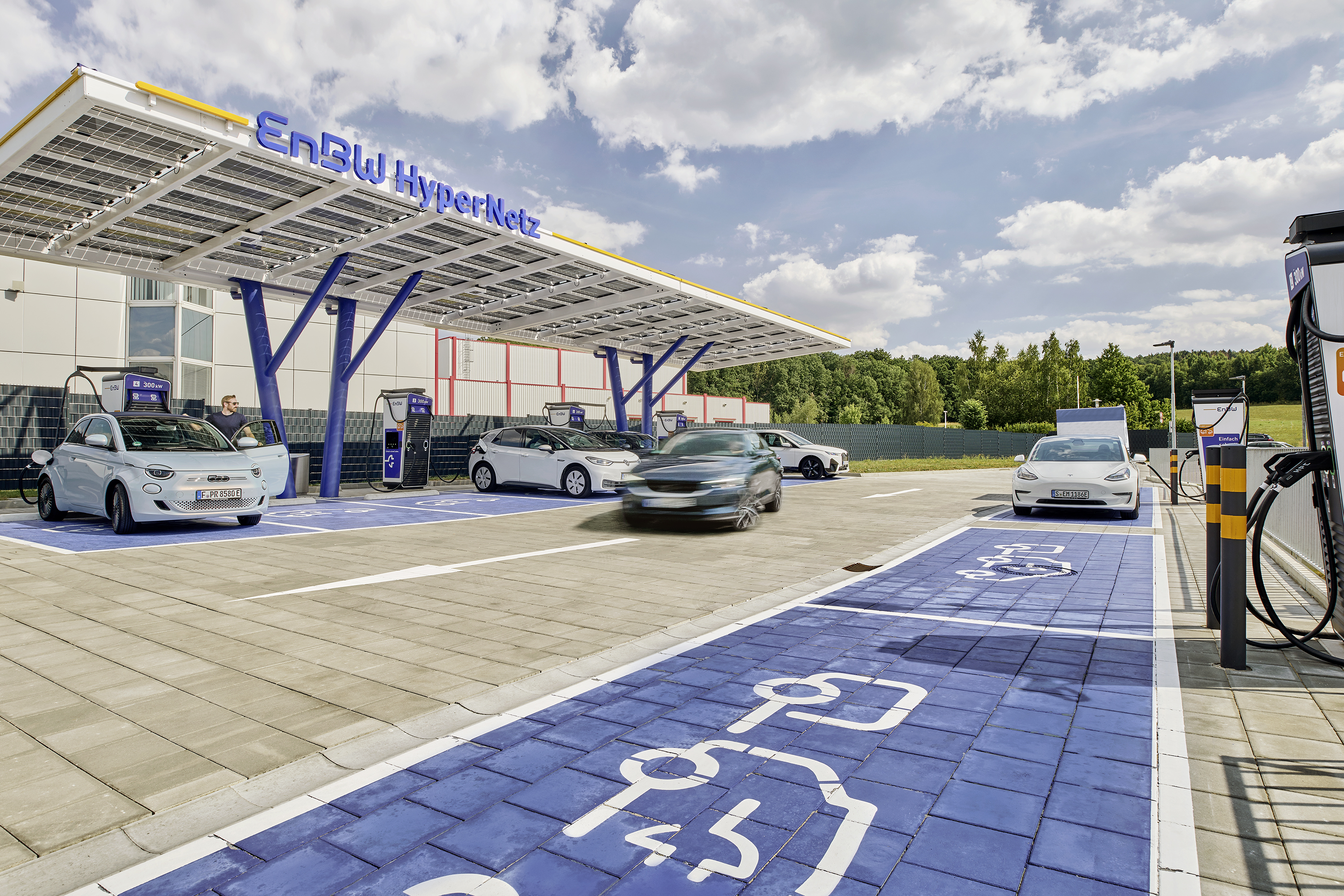 Das Bild zeigt eine moderne Elektroauto-Ladestation mit mehreren Ladepunkten, die von einer weitläufigen Photovoltaikanlage überdacht sind. Verschiedene Elektrofahrzeuge unterschiedlicher Hersteller sind an den Ladepunkten zu sehen, was auf eine breite Nutzung der Anlage hindeutet. Die Gestaltung in kräftigen Blau- und Gelbtönen sowie die hochwertige Architektur verleihen der Ladestation ein technologisch-innovatives Erscheinungsbild.