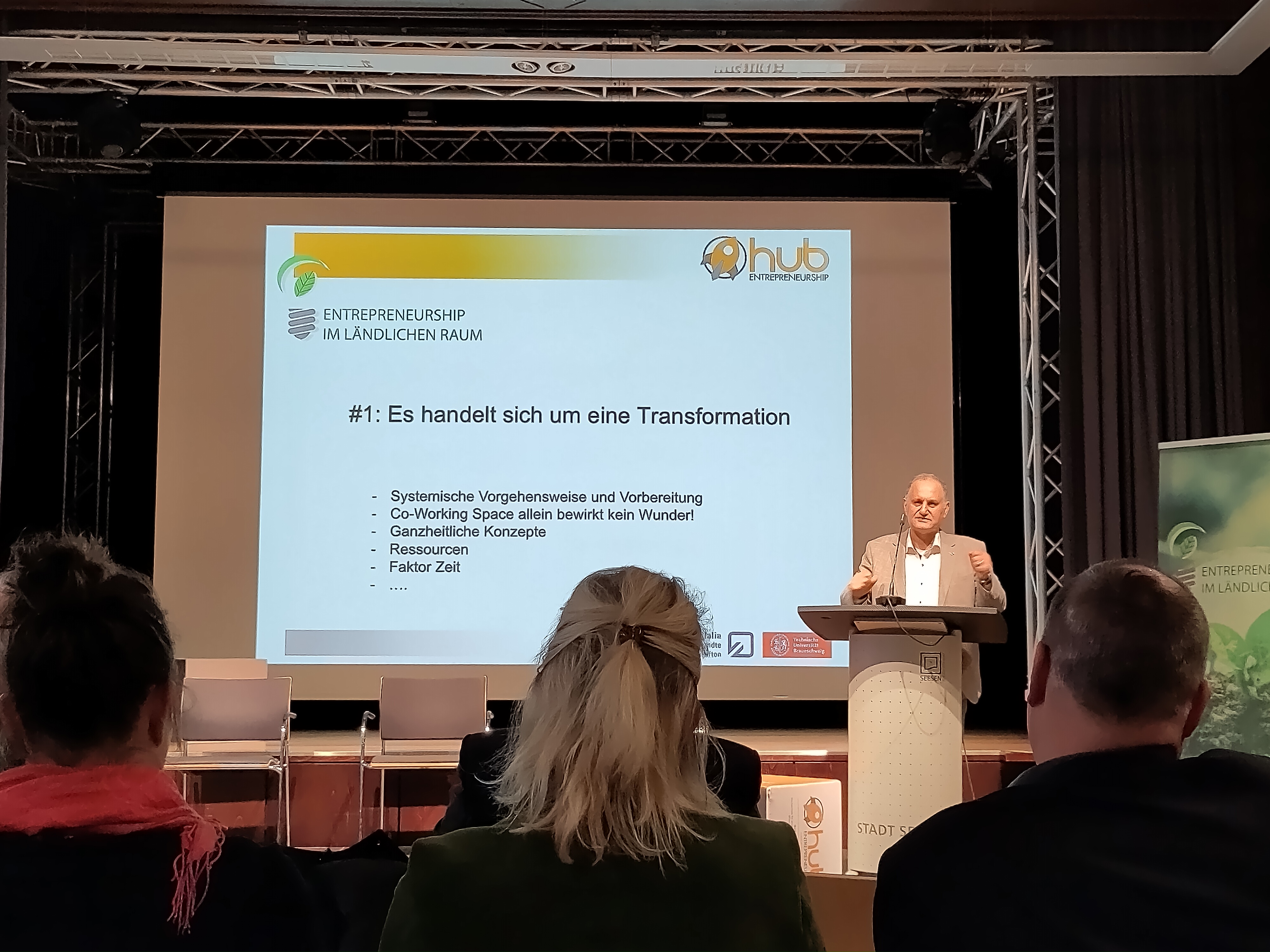 Das Bild zeigt einen Redner auf einer Bühne, der vor einem Publikum einen Vortrag hält. Auf der Leinwand hinter ihm steht der Titel "#1: Es handelt sich um eine Transformation" mit weiteren Stichpunkten zur systemischen Vorgehensweise, ganzheitlichen Konzepten, Ressourcen und dem Faktor Zeit.